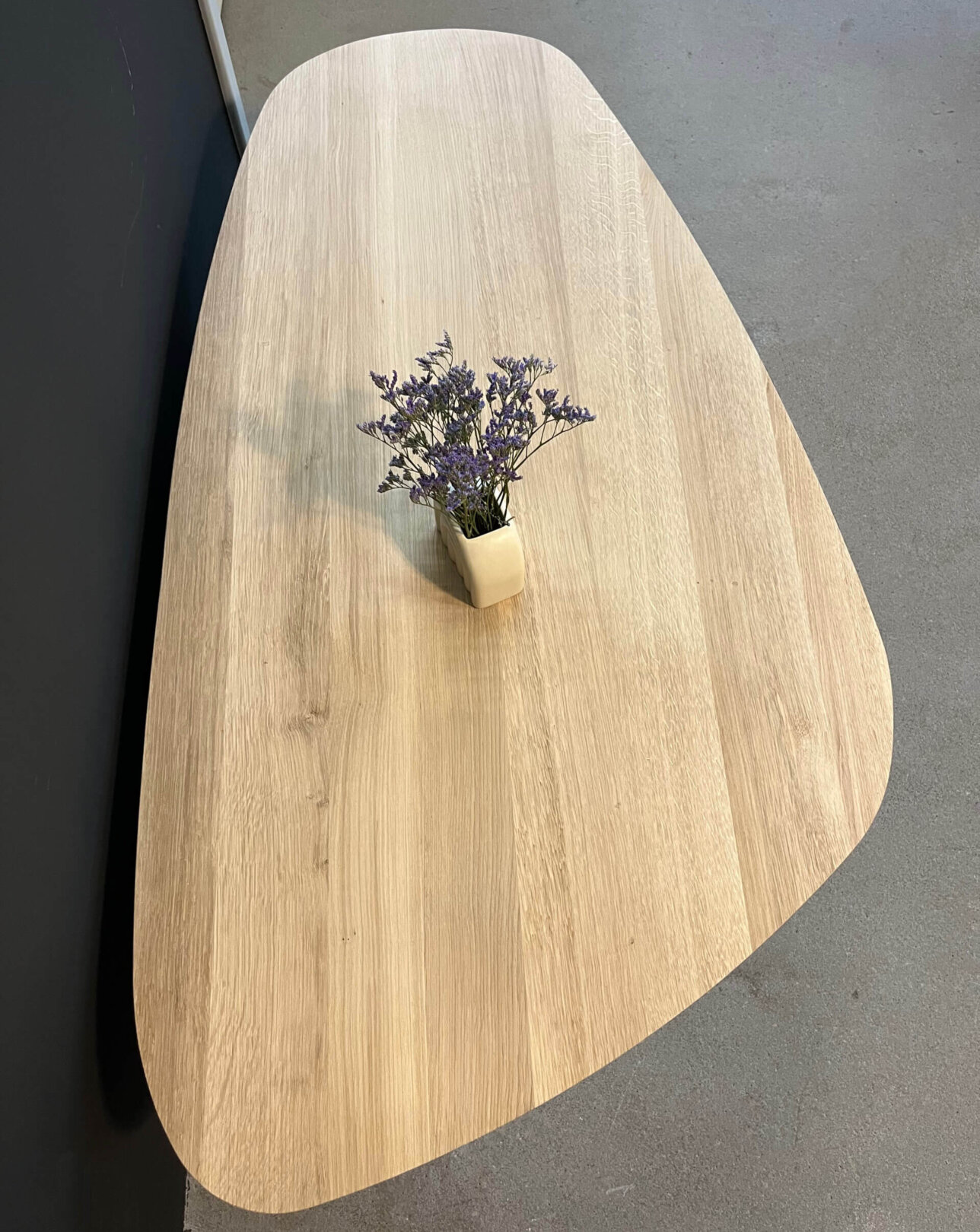 Couchtisch organische form aus heller eiche, kieselform mit schöner maserung, vase mit lavendel. Betonboden und schwarze wand im hintergrund.